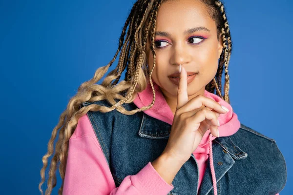 Atractiva mujer afroamericana mostrando shh y mirando hacia otro lado, dedo cerca de los labios, fondo azul - foto de stock