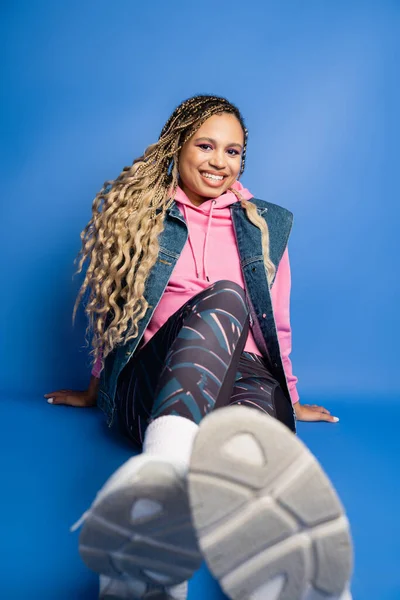 Happy dark skinned woman with dreadlocks sitting in sporty outfit on blue background, look at camera — Stock Photo