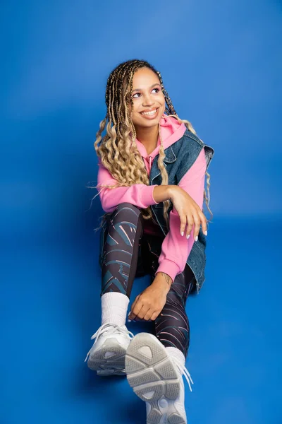 Happy african american woman with dreadlocks sitting on blue background and looking away, sporty — Stock Photo