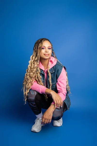 Confident dark skinned woman with dreadlocks sitting in sporty outfit on blue background, smile — Stock Photo