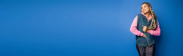 Banner, happy dark skinned woman in denim vest and pink hoodie standing on blue background — Stock Photo