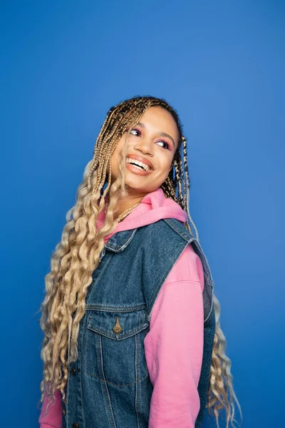 Cheerful african american woman with dreadlocks and bold makeup looking away on blue background — Stock Photo