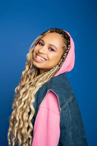 Joyful and pretty african american woman with hood on head smiling on blue background, positive — Stock Photo