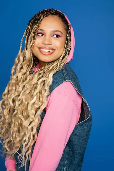 Retrato de mujer afroamericana en capucha rosa sonriendo y mirando hacia otro lado, fondo azul, alegría - foto de stock