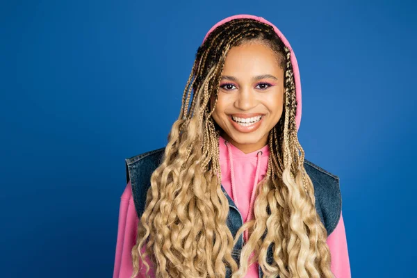 African american woman with bold makeup wearing hoodie and denim vest, smiling in studio on blue — Stock Photo
