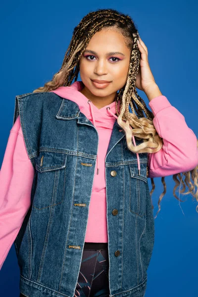 Dark skinned woman with bold makeup smiling on blue, wearing pink hoodie and denim vest in studio — Stock Photo