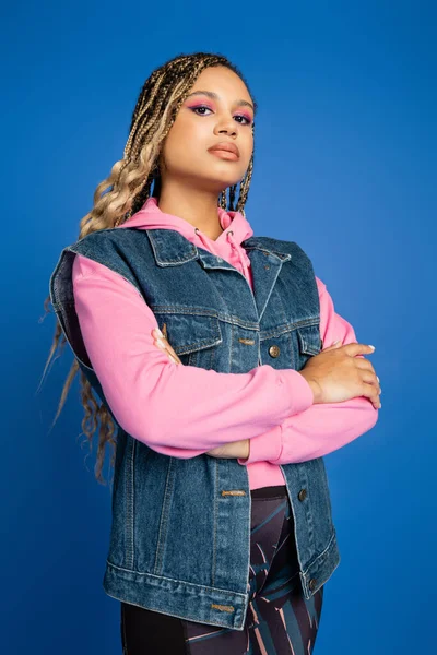 Confident and dark skinned woman standing with folded arms, blue background, denim vest and hoodie — Stock Photo