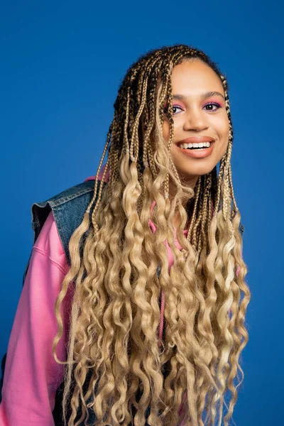 Affascinante donna afro-americana con i capelli lunghi sorridente e guardando la fotocamera su sfondo blu — Foto stock