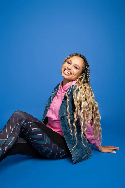 Sporty african american woman with long hair smiling, looking at camera, sitting on blue background — Stock Photo