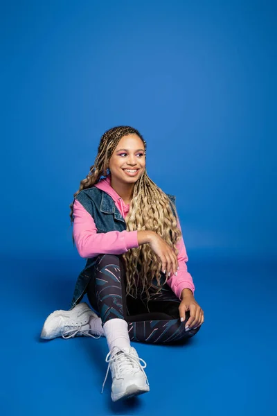 Full length of young long haired african american woman sitting in sporty outfit on blue background — Stock Photo