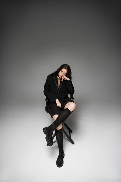Модная молодая азиатка в черной куртке сидит на стуле и позирует на сером фоне — стоковое фото