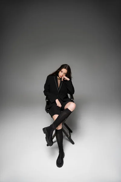 Trendy young asian woman in black jacket sitting on chair and posing on grey background — Stock Photo