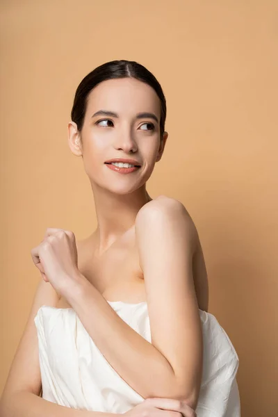 Sonriente joven mujer asiática en la parte superior con los hombros desnudos de pie aislado en beige - foto de stock