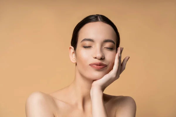 Young asian woman with naked shoulders and closed eyes touching cheek isolated on beige — Stock Photo
