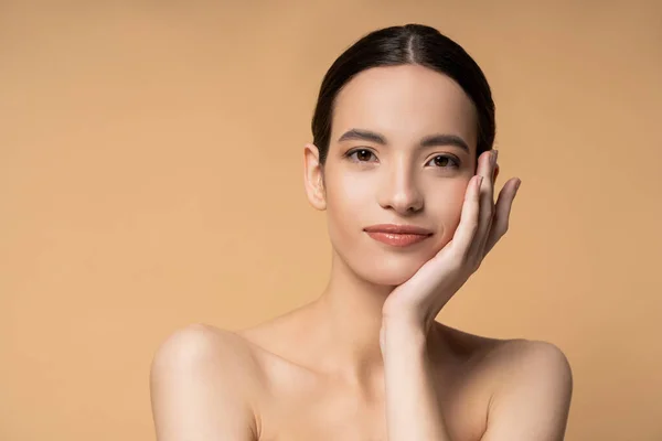 Young asian model with naked shoulders and perfect skin touching cheek isolated on beige — Stock Photo