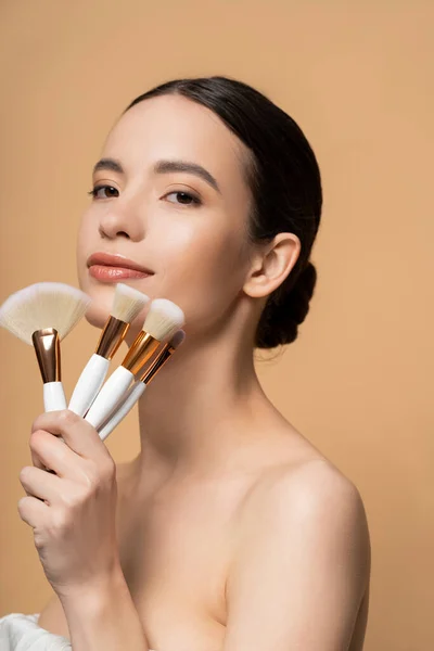 Femme asiatique aux épaules nues tenant des pinceaux de maquillage et regardant la caméra isolée sur beige — Photo de stock