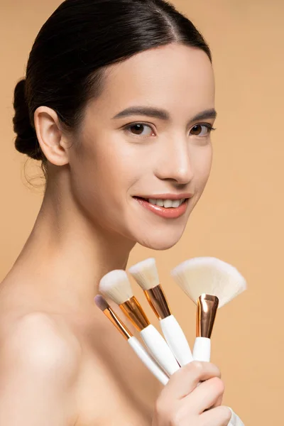 Retrato de modelo asiático sonriente sosteniendo pinceles de maquillaje y mirando a la cámara aislada en beige - foto de stock
