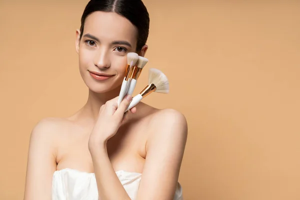 Pretty asian woman with naked shoulders in top holding makeup brushes and smiling isolated on beige — Stock Photo
