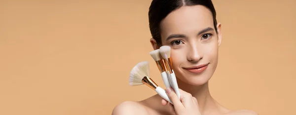 Sonriente modelo asiático con hombros desnudos sosteniendo cepillos de maquillaje aislados en beige, bandera - foto de stock