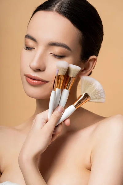 Bella donna asiatica con spalle nude che tengono pennelli per il trucco e gli occhi di chiusura isolati sul beige — Foto stock