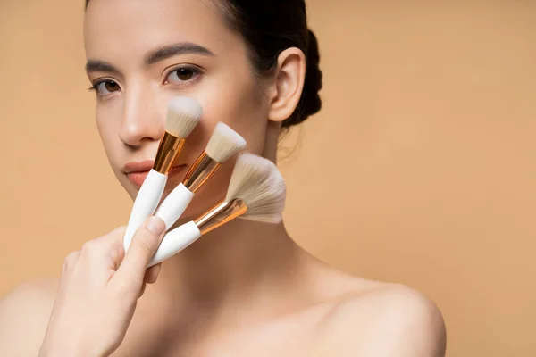 Joven mujer asiática con hombros desnudos sosteniendo cepillos de maquillaje cerca de la cara aislada en beige - foto de stock