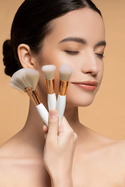 Close up view of young asian woman with perfect skin holding makeup brushes isolated on beige — Stock Photo