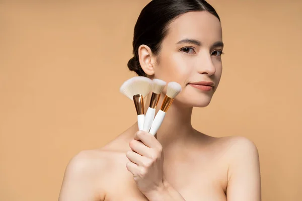 Brunette asiatique femme aux épaules nues tenant des pinceaux de maquillage isolés sur beige — Photo de stock