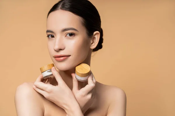 Joven mujer asiática con hombros desnudos sosteniendo frascos con crema cosmética y posando aislado en beige - foto de stock