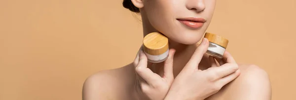 Cropped view of woman with naked shoulders holding jars of cosmetic cream isolated on beige, banner — Stock Photo