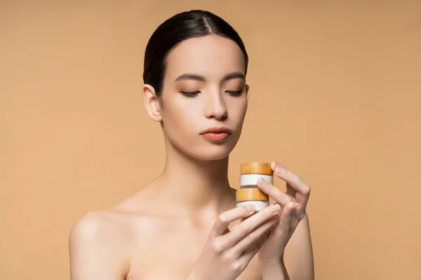 Brunette asian woman with naked shoulders holding jars with cosmetic cream isolated on beige — Stock Photo