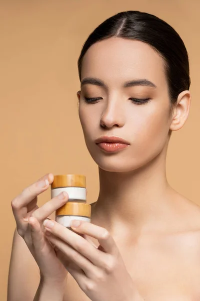 Jovem mulher asiática com ombros nus e pele perfeita segurando creme cosmético isolado no bege — Fotografia de Stock