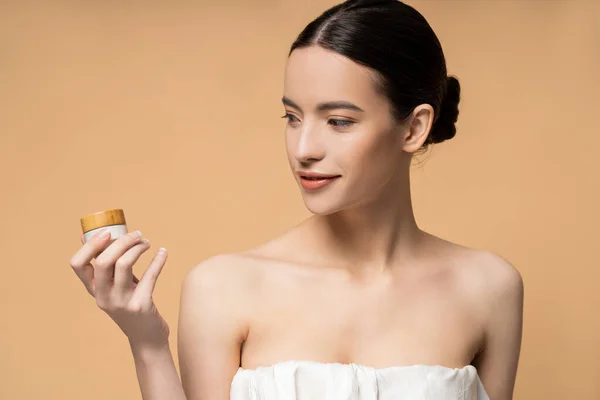 Positivo joven asiático mujer en la parte superior celebración de crema cosmética, mientras que de pie aislado en beige - foto de stock