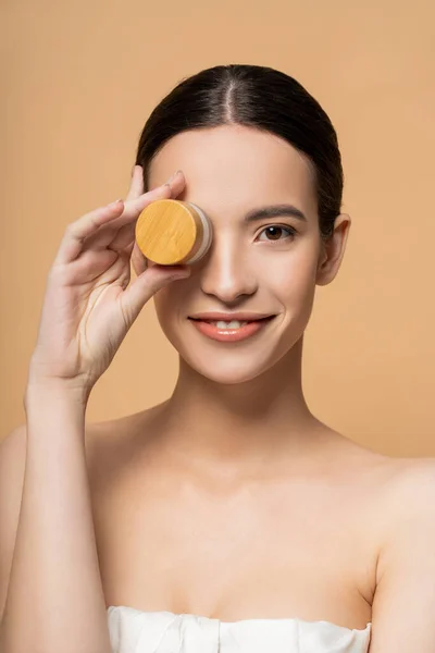Fröhliche asiatische Frau in Top mit nackten Schultern hält kosmetische Creme in der Nähe Gesicht isoliert auf beige — Stockfoto