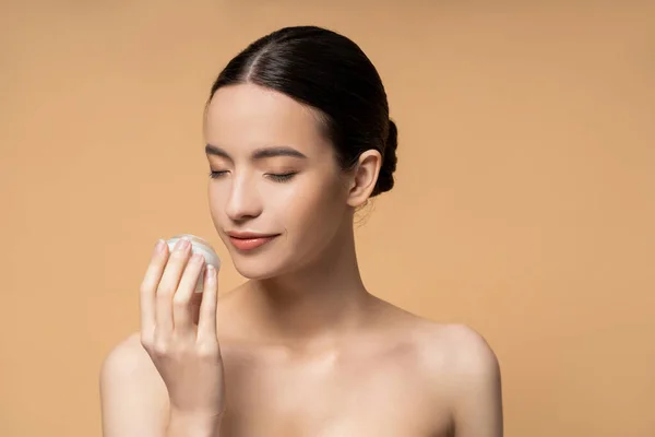 Joven mujer asiática con hombros desnudos oliendo crema cosmética mientras está de pie aislado en beige - foto de stock