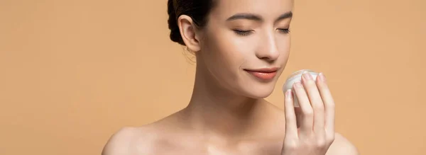 Mujer asiática joven con hombros desnudos oliendo crema cosmética aislada en beige, pancarta - foto de stock