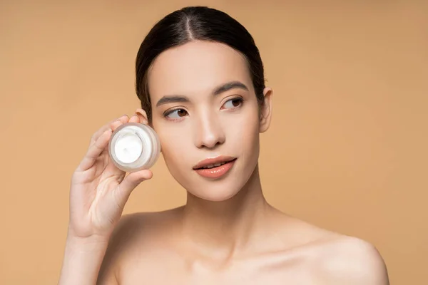 Young brunette woman with perfect skin and naked shoulders holding cosmetic cream isolated on beige — Stock Photo