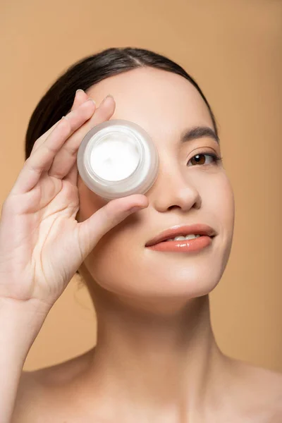 Gioioso giovane donna asiatica in possesso di crema cosmetica vicino al viso e in piedi isolato su beige — Foto stock