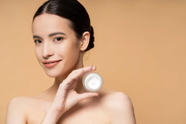 Happy asian woman with naked shoulders and natural makeup holding cosmetic cream isolated on beige — Stock Photo