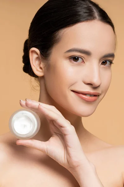 Positiva mujer asiática con la piel perfecta celebración de crema hidratante y mirando a la cámara aislada en beige - foto de stock