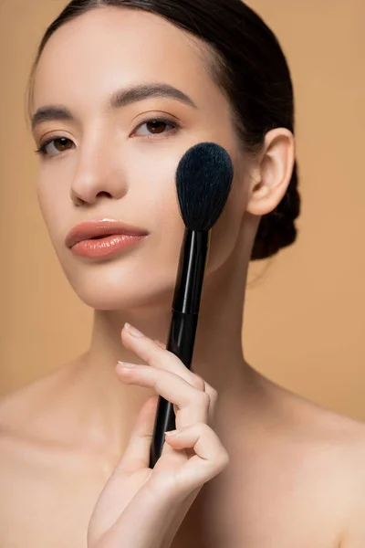 Joven mujer asiática con maquillaje natural y hombros desnudos sosteniendo cepillo rubor aislado en beige - foto de stock