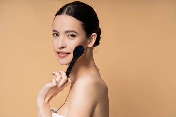 Sorridente giovane donna asiatica con spalle nude tenendo pennello arrossito vicino al viso isolato sul beige — Foto stock