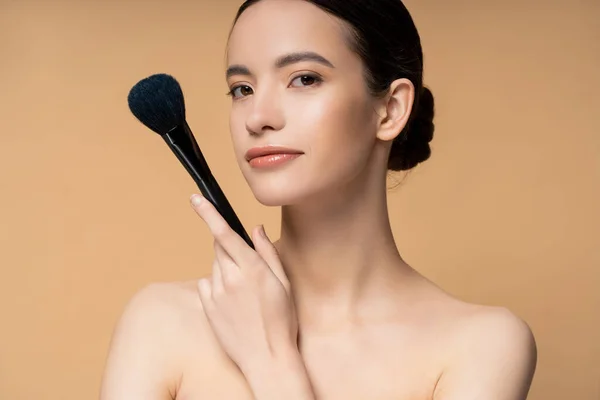 Confiado joven mujer asiática con hombros desnudos sosteniendo cepillo de maquillaje y de pie aislado en beige - foto de stock