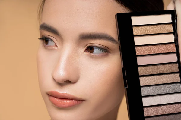 Close up vista de jovem mulher asiática com maquiagem natural segurando paleta de sombra isolada no bege — Fotografia de Stock