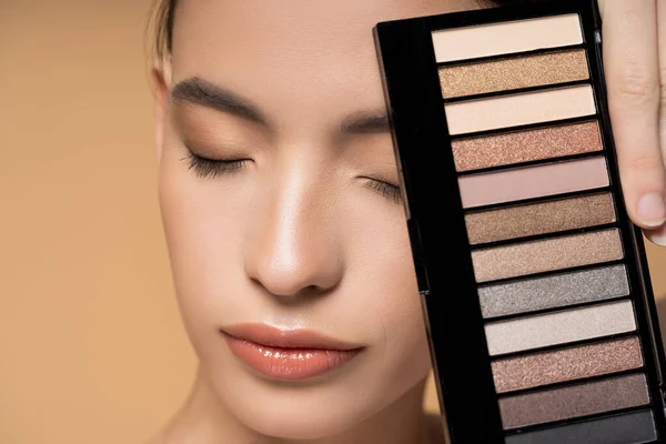 Close up view of asian woman with natural visage holding eyeshadow palette isolated on beige — Stock Photo