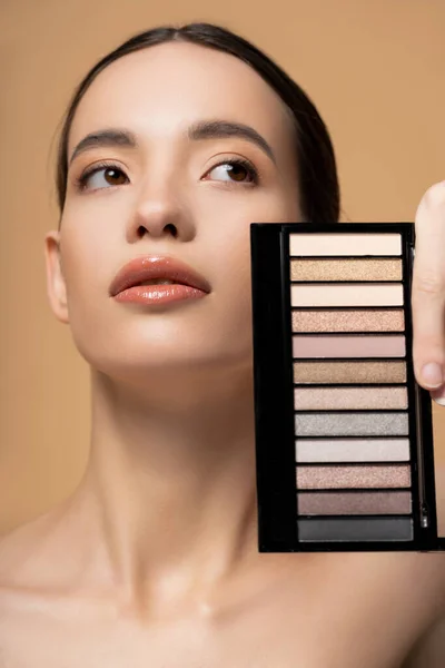 Jeune femme asiatique avec maquillage naturel tenant palette de fards à paupières et regardant loin isolé sur beige — Photo de stock