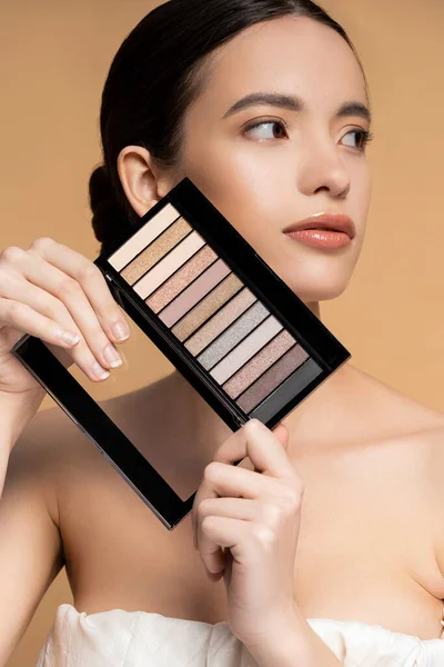 Asian model with perfect skin holding eyeshadow makeup palette while posing isolated on beige — Stock Photo