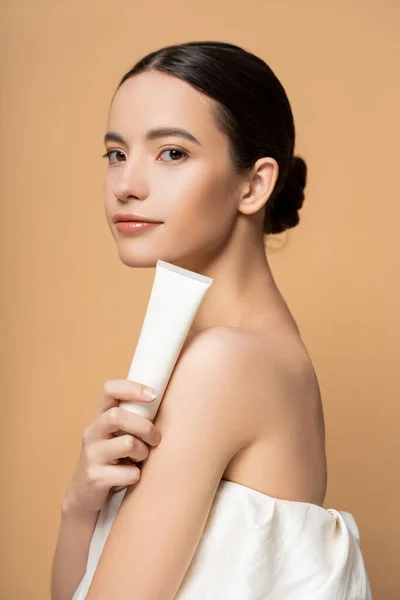 Pretty young asian model in top holding cosmetic lotion and looking at camera isolated on beige — Stock Photo