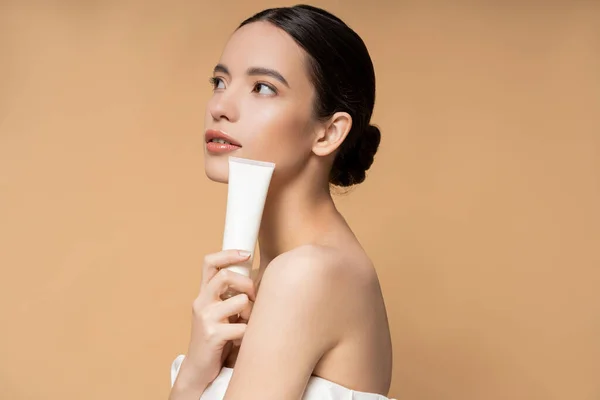 Young asian woman in top holding cosmetic lotion and looking away isolated on beige — Stock Photo