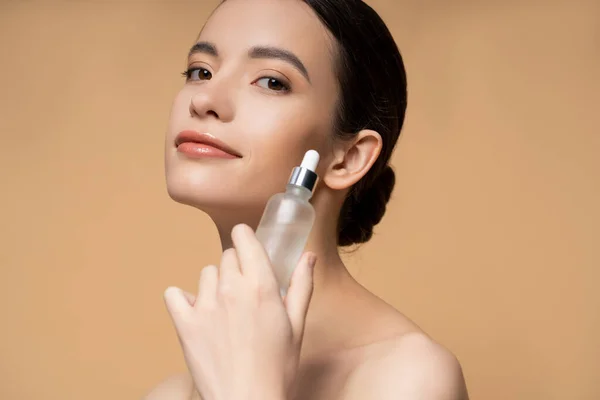 Pleased young asian woman with naked shoulders holding cosmetic serum isolated on beige — Stock Photo