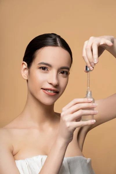 Positive young asian woman with naked shoulders holding cosmetic serum isolated on beige — Stock Photo